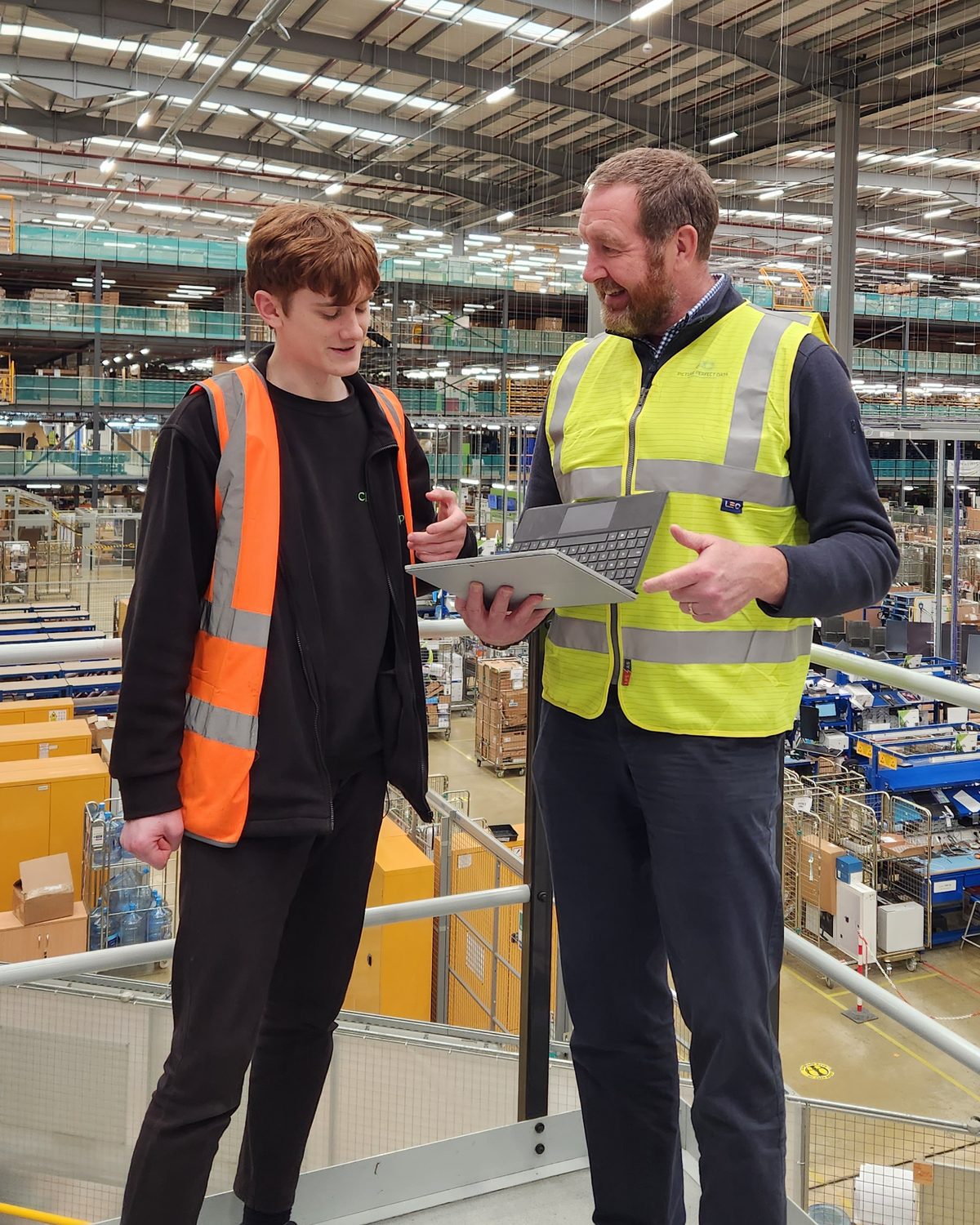 Steve Coleman and a colleague in hi-vis on a mezzanine above a busy warehouse floor, looking at a laptop together